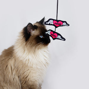 Cat sitting and facing the right with a dangly toy in front of its face that looks like two hearts with wings. "Fucks" is written on the heart. Dangly cat toy with speech bubble that says, "Zero", connected to two hearts with wings and "Fucks" written on them. An original from the Lucky Bubs pet store.